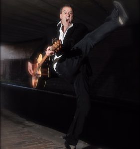 Comedian Boothby Graffoe kicking his leg in an underpass with his guitar