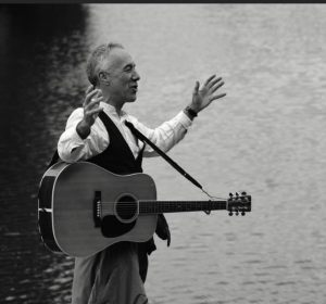 Ray HEarne in black and white with his guitar in front of water