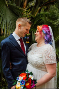 Ellie and David in wedding clothes in front of green plants, Ellie looks a little better than David, which is only to be expected, but he does look smashing. Photo by Ellie Grace Photography